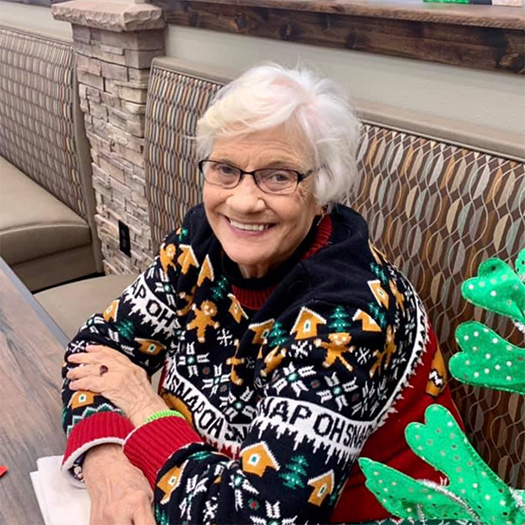 An elderly woman in a Christmas sweater sitting at a table.