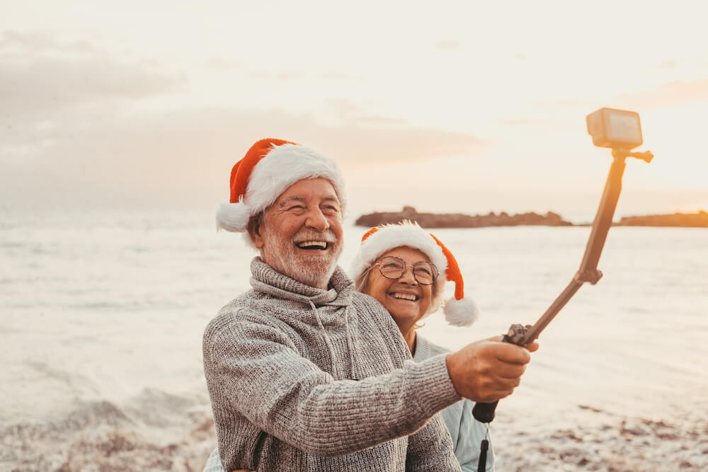 An elderly couple in Santa hats taking a selfie on the beach.