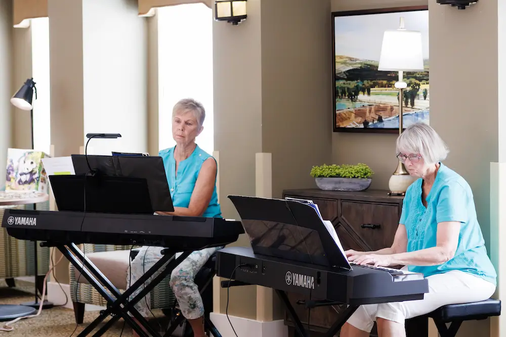 Two elderly women in blue tops sit at Yamaha electronic pianos and play music in a bright, well-lit living room.