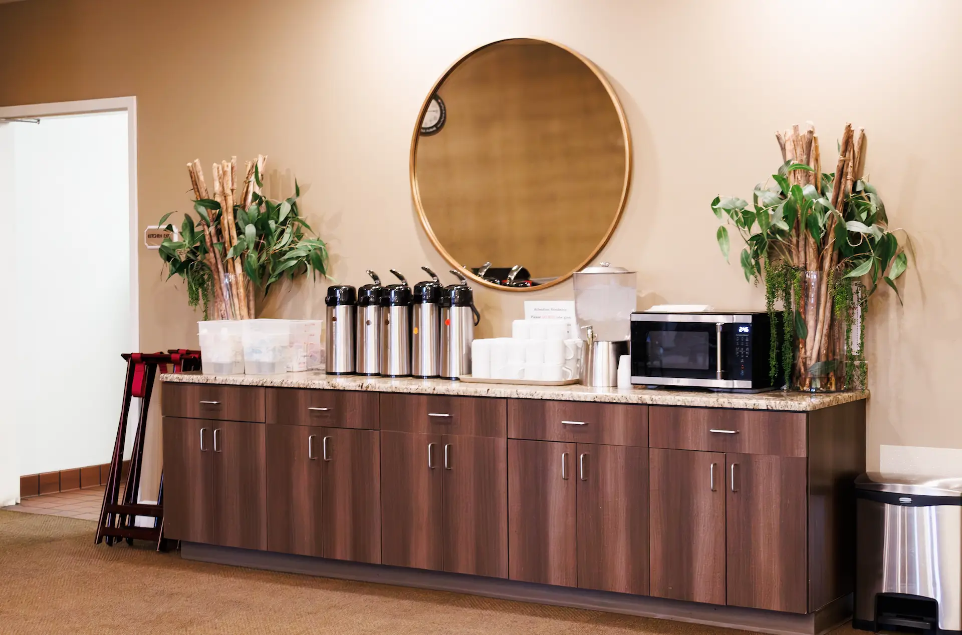 A long wooden beverage station in a conference room with a round wall mirror, bottled drinks and a microwave on the counter, and plants on both ends.