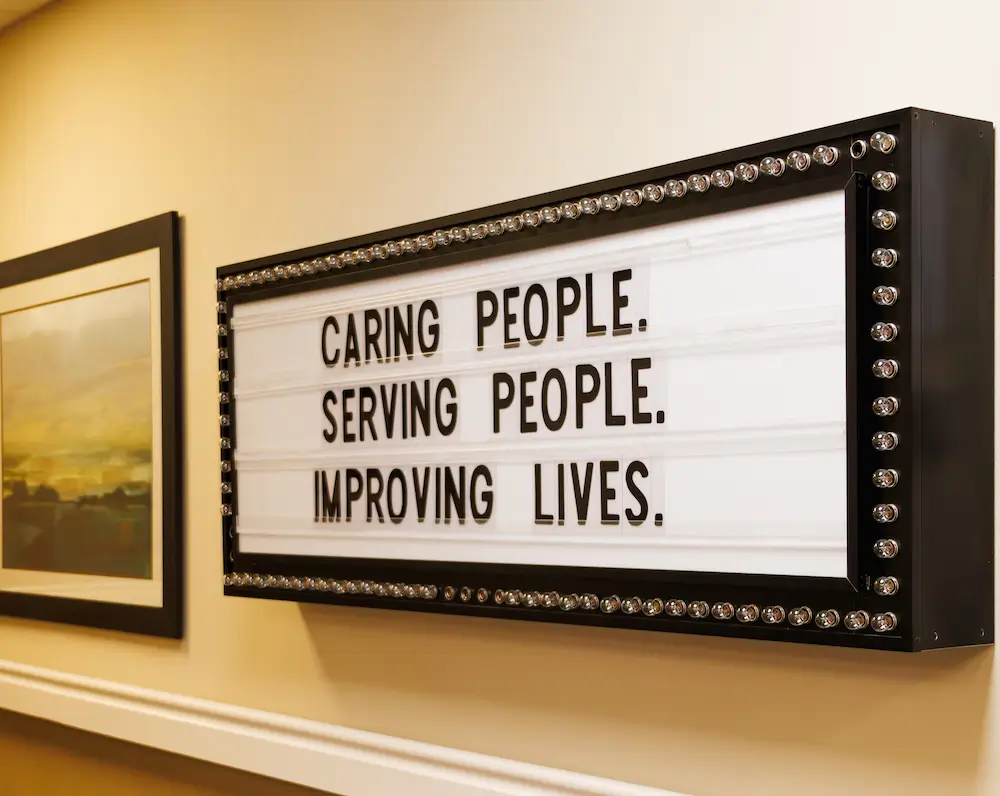 Framed lightbox on a wall displaying the motto "Caring people. Serving people. Improving lives."
