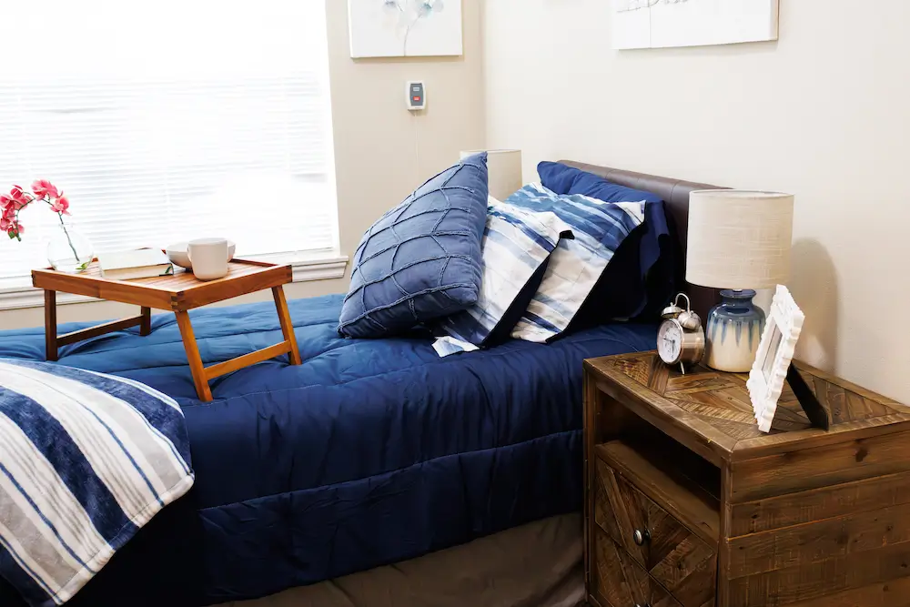 Cozy bedroom with a navy blue bedspread and pillows, a wooden nightstand with a lamp, and a bed tray holding a cup.