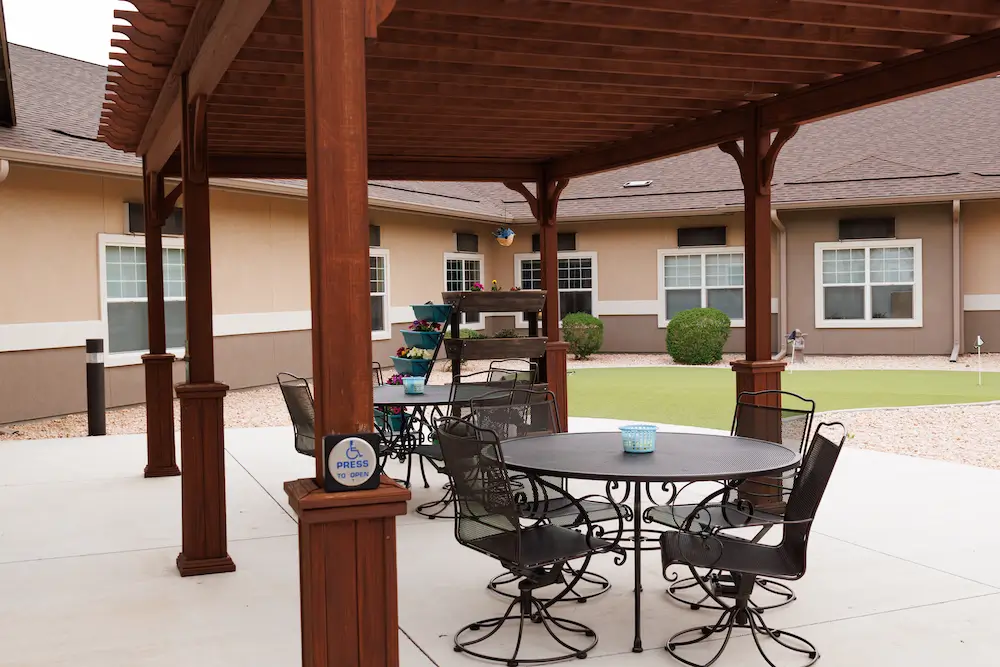 Outdoor patio under a wooden pergola with a round metal table and chairs, near a beige apartment building.