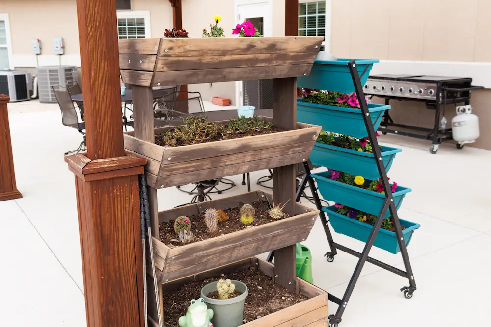 Weathered wooden multi-tier planter rack filled with small potted succulents sits on a bright patio beside a blue rolling plant stand, with a grill and outdoor kitchen area in the background.