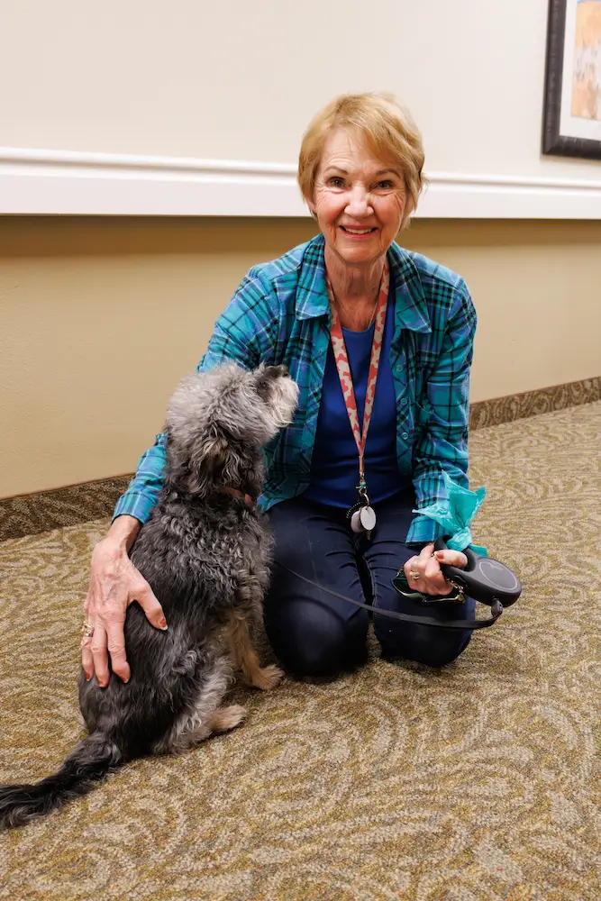 An elderly woman sits on a carpeted floor, petting a small gray dog.