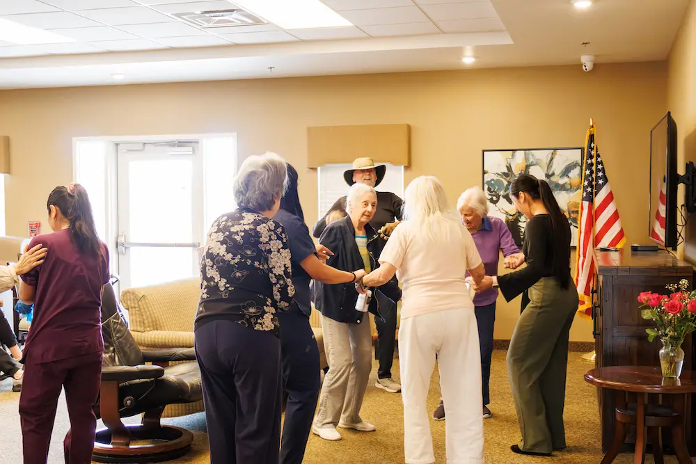A group of people stand in a circle in a warmly lit room, holding hands around a man wearing a cowboy hat with an American flag in the background.