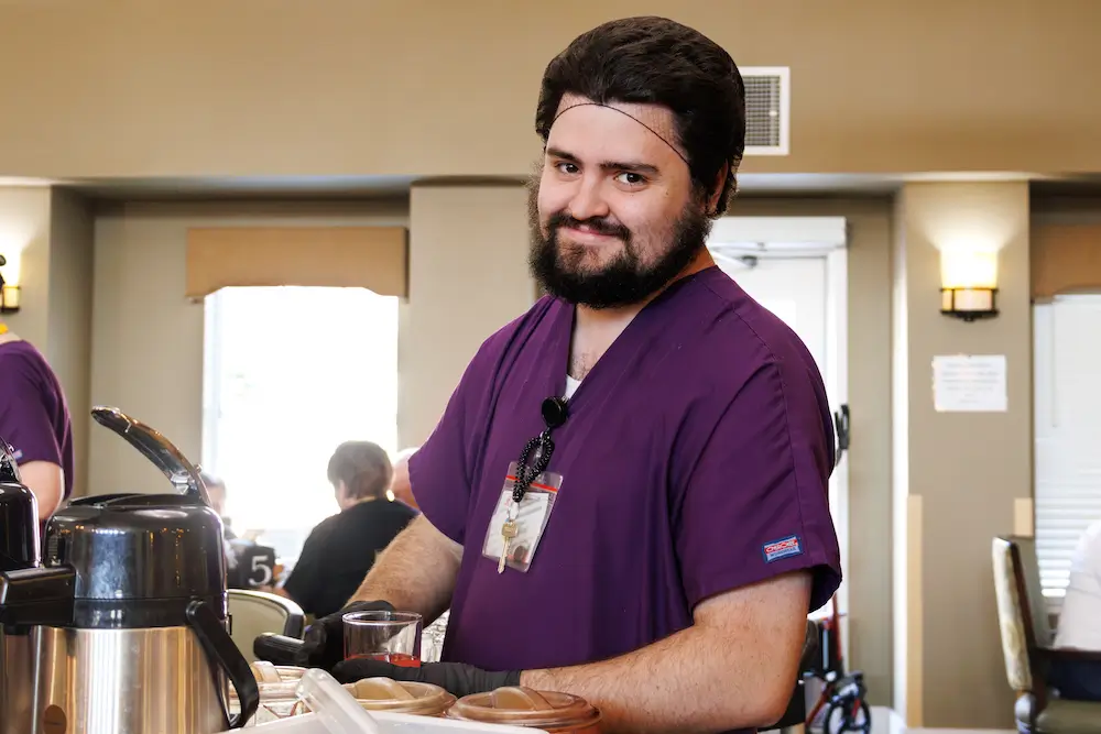 Smiling man in purple scrubs with a badge stands behind a coffee station.