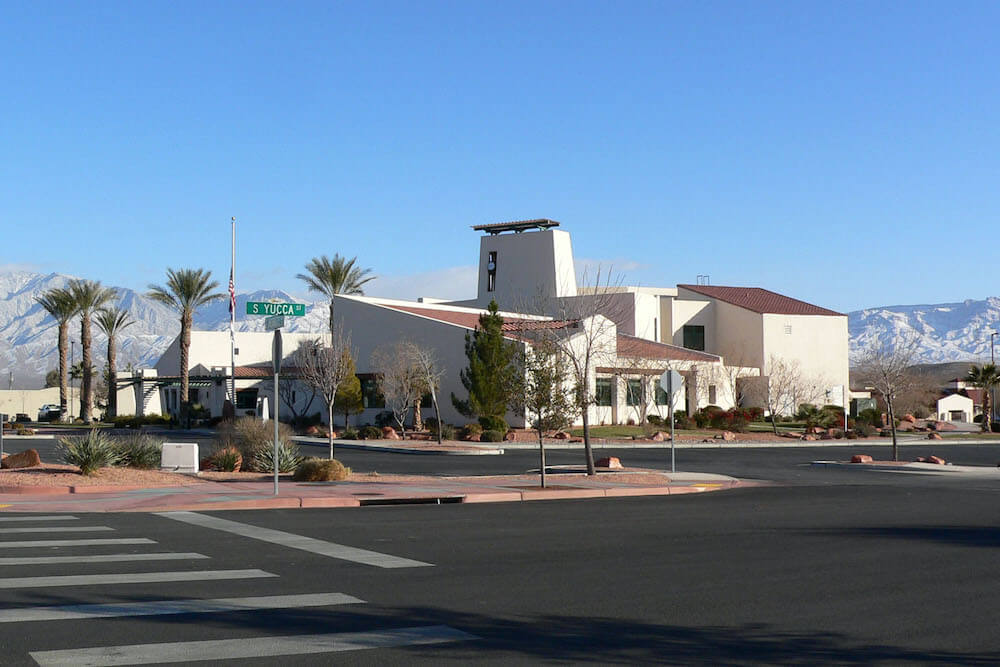 Modern church building with bell tower, surrounded by palm trees and mountains. Intersection with street sign in foreground, clear blue sky above.