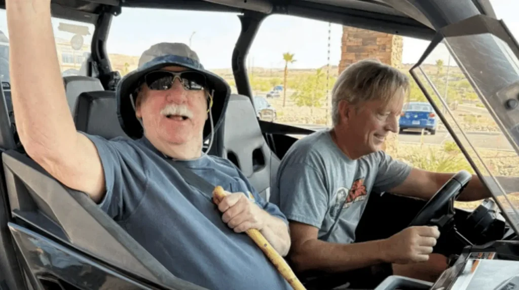 Two men sit in a race car cockpit, one wearing a hat and sunglasses raising his arm in excitement while the other smiles and drives.