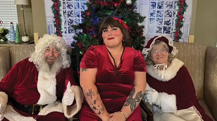A woman in a red dress sits between two people dressed as Santa Claus on a sofa in a living room with a decorated Christmas tree.