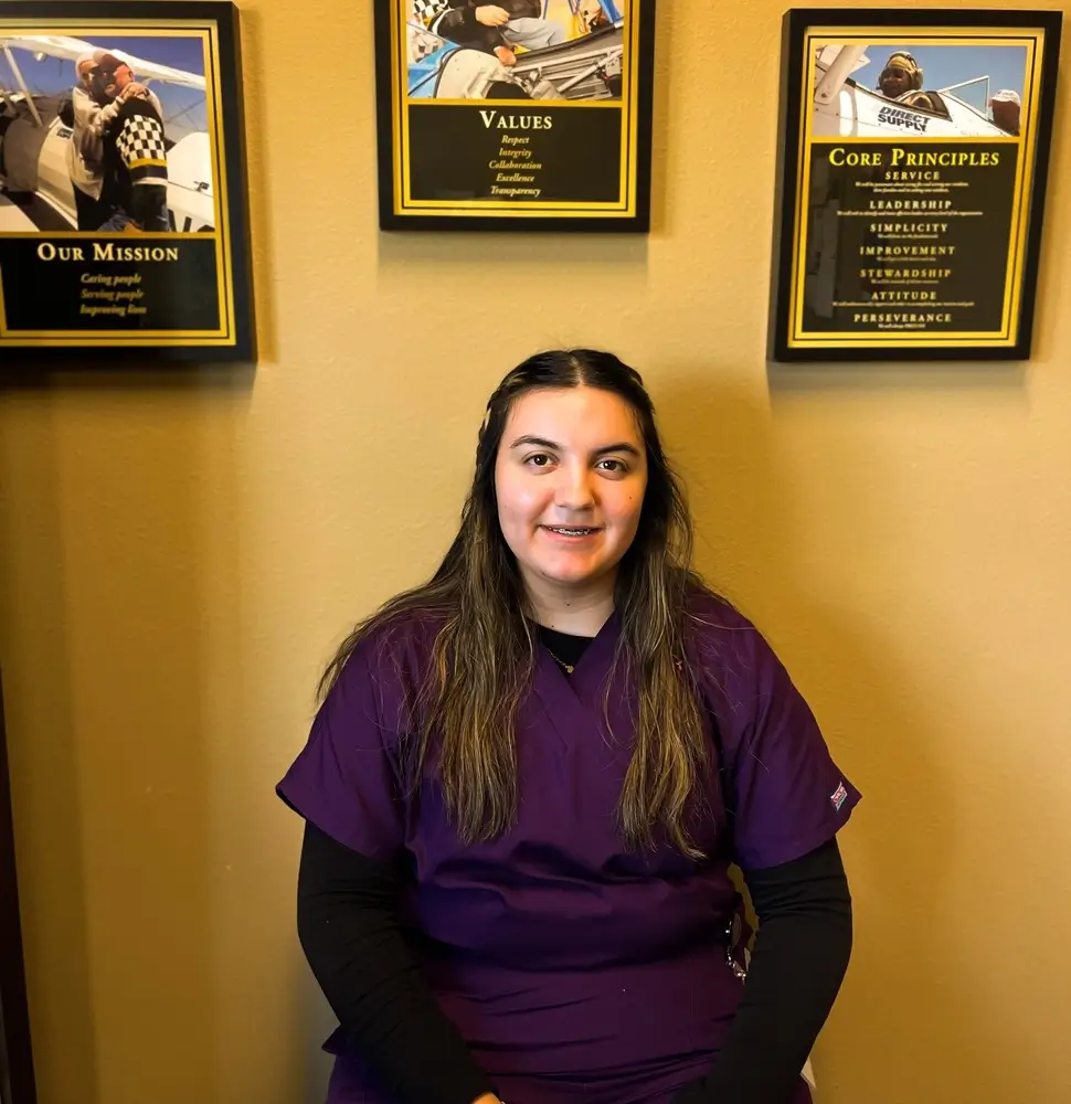 Woman in purple scrubs sits in front of a yellow wall with framed posters labeled Our Mission, Values, and Core Principles.