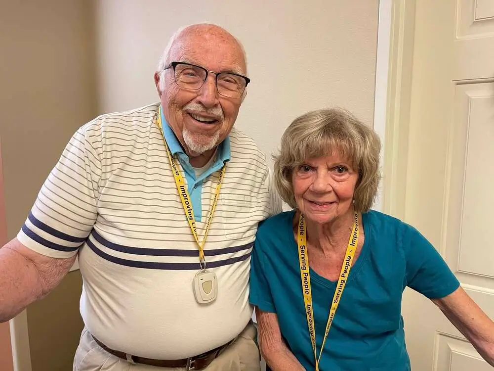 An elderly man and woman stand together indoors, smiling, each wearing a yellow lanyard with a badge.