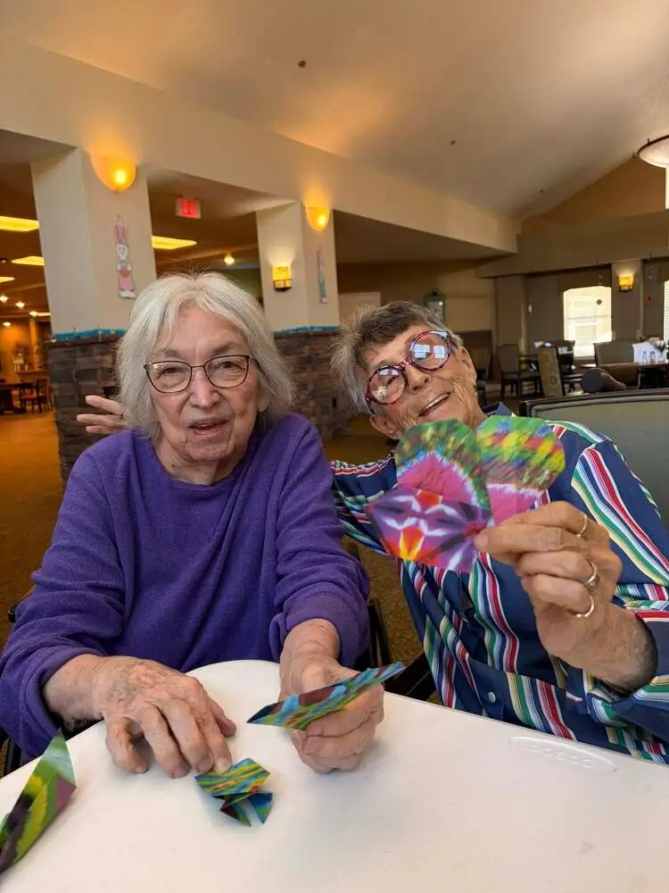 Two elderly women sit at a bright table, proudly displaying colorful folded paper crafts.