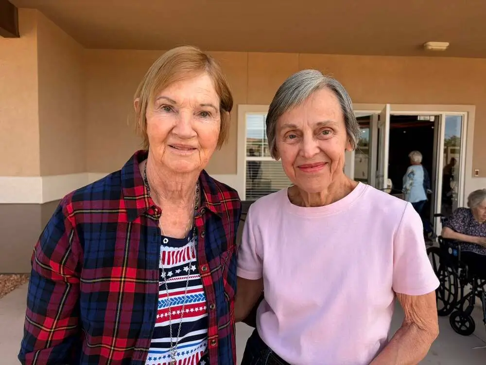 Two elderly women stand together outside a building, smiling at the camera; one wears a red plaid shirt and the other a light pink top.