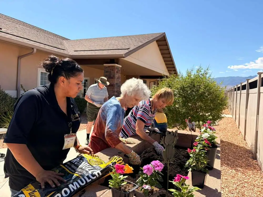 A group of volunteers gardening outside a house, planting colorful flowers in pots along a gravel path.
