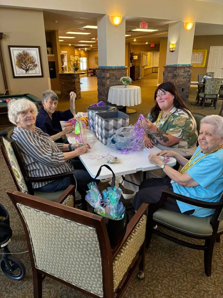 Four elderly people sit around a table in a senior living facility, with gift bags and purple tissue paper as they celebrate.