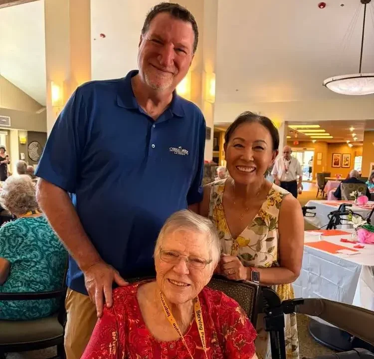 Three smiling people pose together at a social event, with an elderly woman in a wheelchair seated in front.