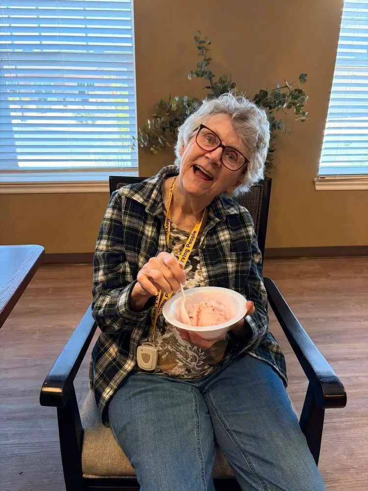 Smiling elderly woman sitting in a chair, wearing glasses and a plaid shirt, eating from a bowl with a spoon.
