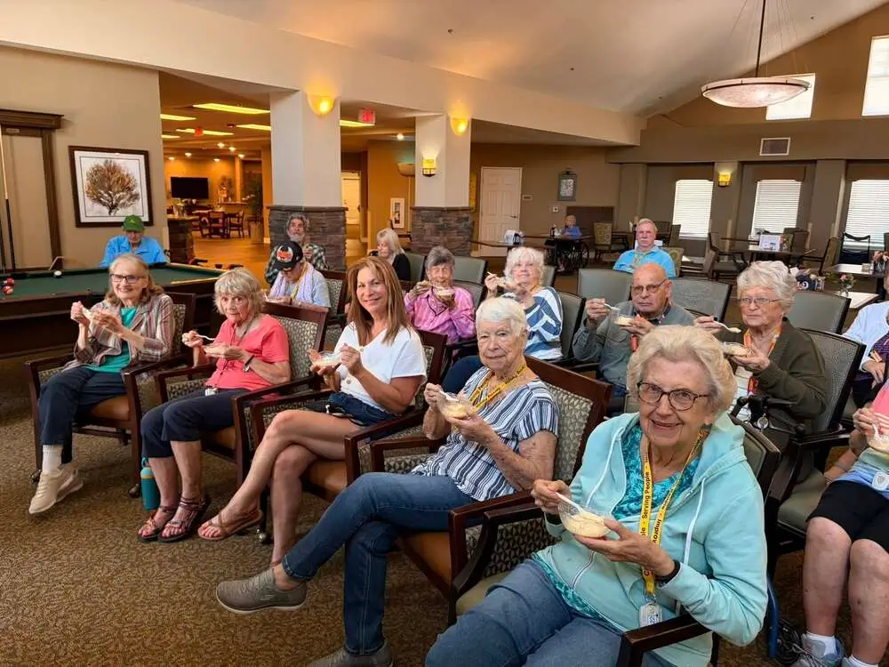 A group of elderly residents seated in a bright lounge, with some reading newspapers.
