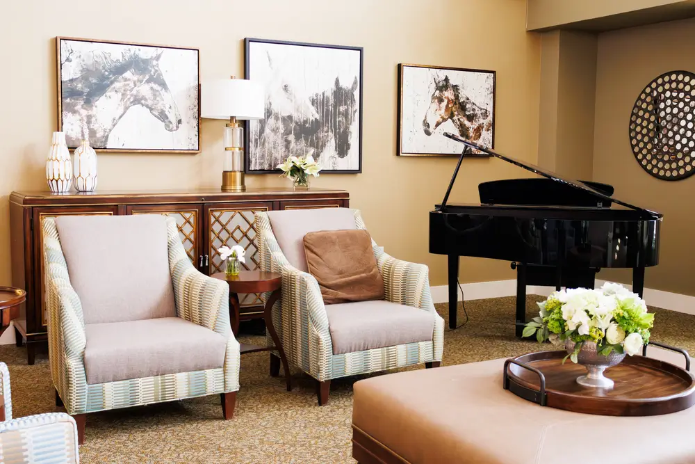 A cozy living room with two upholstered armchairs, a grand piano, framed wall art, and a round coffee table topped with a flower arrangement.