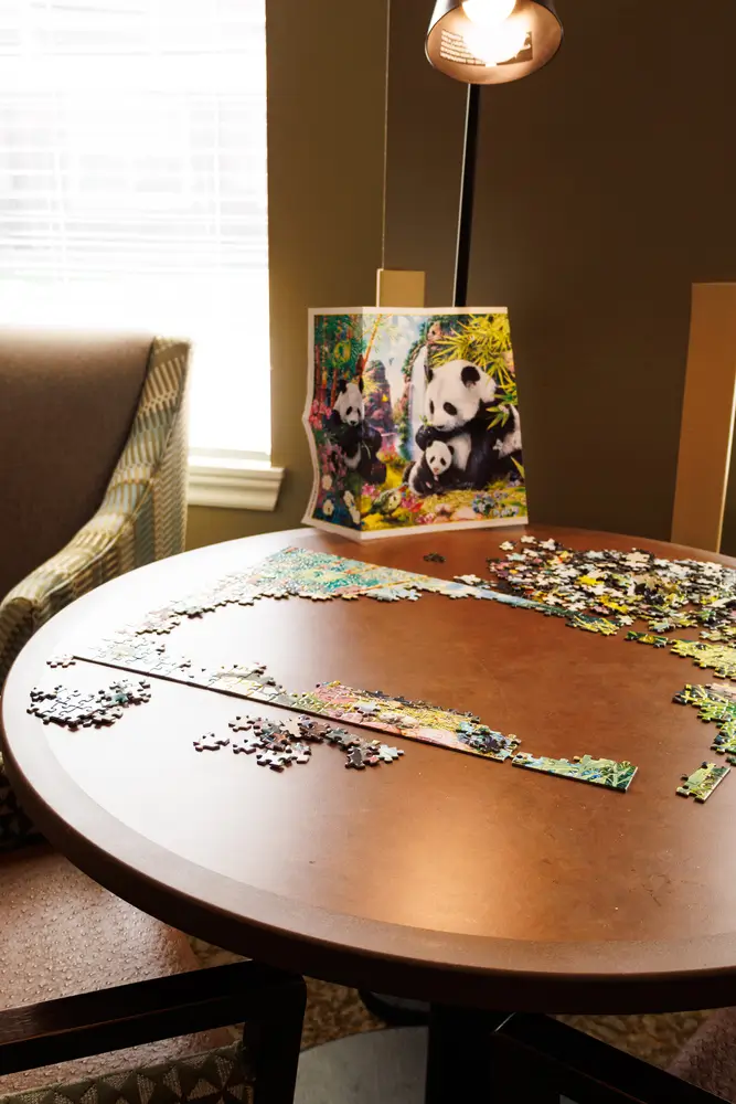 A round wooden table with scattered jigsaw puzzle pieces, a panda painting leaning against the wall behind it, and a standing lamp in a cozy room.