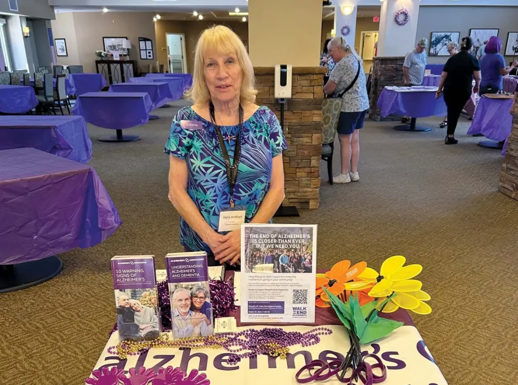 A woman in a blue patterned top stands behind a promotional table with brochures and colorful paper flowers at an indoor convention.