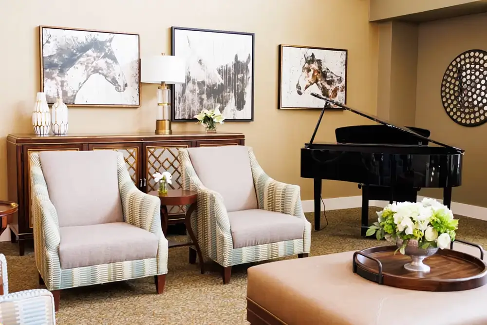 A stylish living room featuring two light upholstered armchairs facing a round coffee table with a floral centerpiece, a grand piano, and framed art on beige walls.