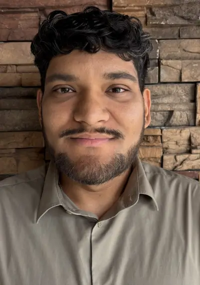 A man with short dark hair and a light beard, wearing a light gray collared shirt, smiles softly in front of a stone wall.